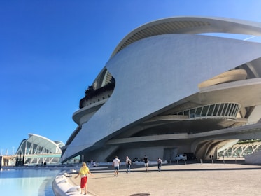 A futuristic architectural structure with sweeping curves and intricate lines dominates the scene. The building features large windows and terraces and is made of concrete and glass. In the foreground, a bright blue sky stretches overhead, and several people walk along a wide promenade near a pool of water.