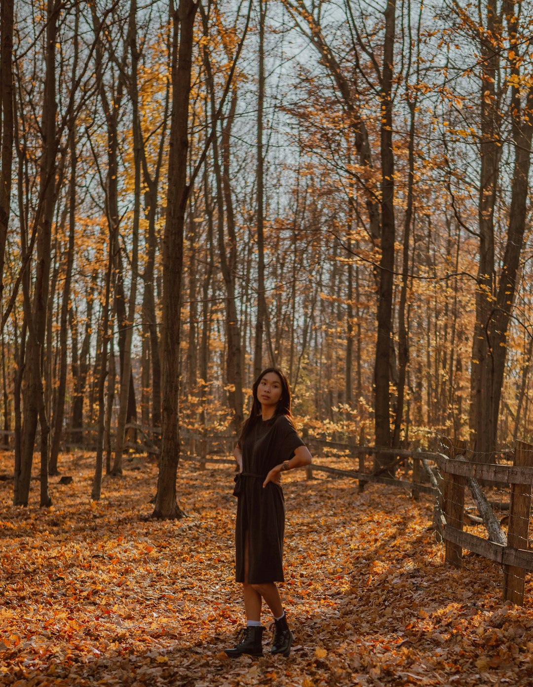 woman in black coat standing on brown dried leaves on ground surrounded by trees during daytime