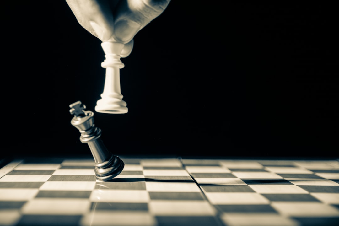 grayscale photo of person holding glass, Close up of white king taking down black king. Hand and fingers and chess board with vintage look