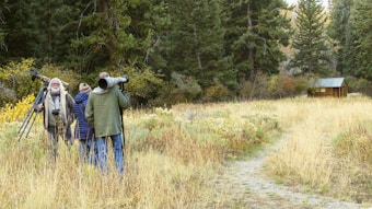 A group of people stand in a grassy field surrounded by dense trees. They are holding cameras with large lenses and tripods, suggesting they are wildlife photographers. The scene is set in a natural environment with a small wooden cabin visible in the background.