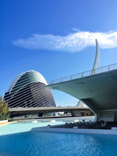 A modern architectural scene featuring a futuristic building with a large, oval-shaped dome, partially covered in a grid-like structure. The building is set against a bright blue sky with some white clouds. In the foreground, a bridge with a sleek, curved design extends across a pool of clear blue water. Several bicycles are parked under the bridge, adding an element of leisure to the scene.