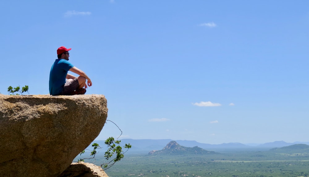 man in blue t-shirt sitting on brown rock near body of water during daytime