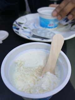 A close-up of a cup containing vanilla ice cream with a wooden spoon inside. In the background, another hand is holding a similar cup, placed on a decorative plate.