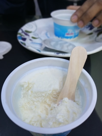 A close-up of a cup containing vanilla ice cream with a wooden spoon inside. In the background, another hand is holding a similar cup, placed on a decorative plate.