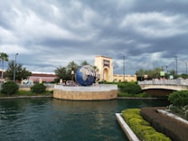 A large globe structure prominently displays the word 'UNIVERSAL' near the entrance of a theme park, surrounded by lush greenery and a bridge leading into the park. The sky is overcast with dark clouds, adding a dramatic backdrop to the scene.