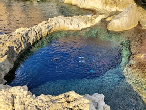 A natural rock pool with clear blue water is surrounded by jagged rock formations. The pool is connected to the sea, and a person is snorkeling in the water, visible by their snorkel and fins. Sunlight casts reflections on the water, highlighting different shades of blue and green.