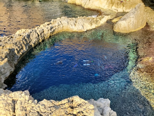 A natural rock pool with clear blue water is surrounded by jagged rock formations. The pool is connected to the sea, and a person is snorkeling in the water, visible by their snorkel and fins. Sunlight casts reflections on the water, highlighting different shades of blue and green.