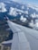 Aerial view capturing the wing of an airplane in flight, with a landscape below consisting of lush greenery, a coastline, and a vast expanse of ocean. The sky is scattered with fluffy white clouds, creating a serene and picturesque scene.