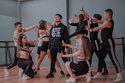A group of dancers is performing in a studio setting. The central figure wears a black hoodie with bold white lettering, surrounded by others in athletic attire, gesturing dynamically. The studio features wooden floors and mirrored walls.