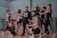 A group of dancers is performing in a studio setting. The central figure wears a black hoodie with bold white lettering, surrounded by others in athletic attire, gesturing dynamically. The studio features wooden floors and mirrored walls.