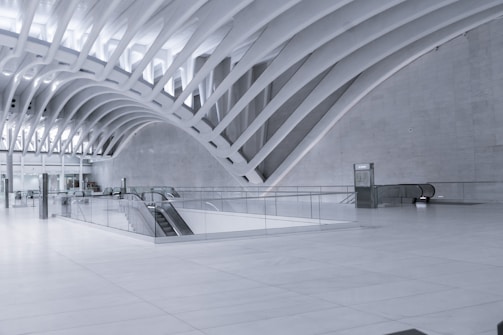 A spacious, modern interior with sweeping white architectural curves and clean lines. The design features a large open area with multiple escalators, a curved ceiling with rib-like beams, and minimalistic detailing.