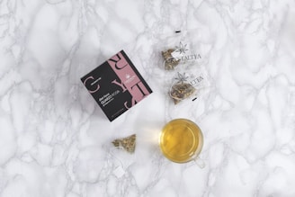 A flat lay composition featuring a box with a black and pink design labeled 'Crystaltea' alongside two clear plastic bags containing herbal tea. There is also a small, transparent teacup filled with light yellow tea and a tea bag with visible herbs near the cup. The background is a white marble surface.