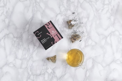 A flat lay composition featuring a box with a black and pink design labeled 'Crystaltea' alongside two clear plastic bags containing herbal tea. There is also a small, transparent teacup filled with light yellow tea and a tea bag with visible herbs near the cup. The background is a white marble surface.