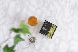A cup filled with amber-colored tea sits on a light marbled surface. Next to it, there is a green and black box labeled Elixir Slim Amazonite High Quality CBD. A blurred green plant is in the foreground, and a small clear bag with a logo is nearby.