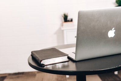 A laptop with a visible Apple logo rests on a round black table next to a closed black book. In the background, there is a white wall and a small white cabinet with a plant on top.