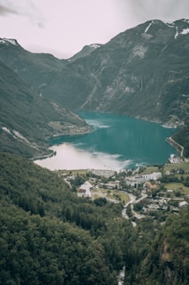 A picturesque fjord landscape with steep, lush green mountains surrounding a serene turquoise body of water. A small village with buildings and a white church is nestled at the foot of the mountains, surrounded by dense forest. The sky is overcast, adding a calm yet dramatic atmosphere to the scene.