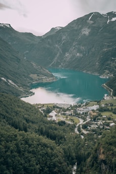 A picturesque fjord landscape with steep, lush green mountains surrounding a serene turquoise body of water. A small village with buildings and a white church is nestled at the foot of the mountains, surrounded by dense forest. The sky is overcast, adding a calm yet dramatic atmosphere to the scene.