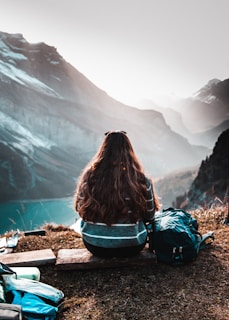 A person with long hair is sitting on a wooden plank overlooking a scenic mountainous landscape. The view includes steep cliffs and a mist-filled valley with a blue body of water in the distance. A backpack and other gear are placed nearby, suggesting a hiking or outdoor excursion.