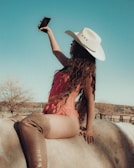 A woman wearing a white cowboy hat and a pink outfit is sitting on a horse while taking a selfie with her smartphone. Her long hair cascades down her back, and she is wearing thigh-high brown boots. The backdrop features a clear blue sky and a sparse rural landscape with bare trees.