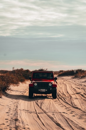 A red Jeep is driving off-road on a sandy beach path. The vehicle is surrounded by sparse vegetation and under a cloudy sky, creating tracks in the sand. There is a signpost visible to the right.