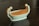 An elegant ceramic dish or bowl, shaped like a boat, adorned with two sculpted figures standing at either end. The figures are gracefully posed and appear to be female, each bowing slightly towards the center. The overall texture is smooth, and the finish is a creamy off-white color.