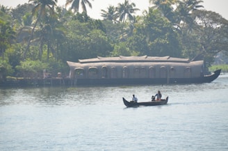 A serene waterway scene featuring a traditional houseboat in the background with lush green trees and palm trees along the shoreline. In the foreground, a small canoe with three people is gently moving on the calm water.