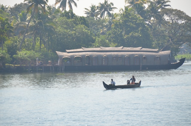 A serene waterway scene featuring a traditional houseboat in the background with lush green trees and palm trees along the shoreline. In the foreground, a small canoe with three people is gently moving on the calm water.