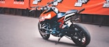 A sporty motorcycle with an orange and black color scheme is parked in front of a backdrop featuring branded banners. The design includes sharp angles and a modern aesthetic, suitable for racing or performance riding.