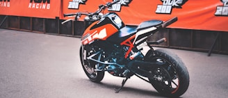 A sporty motorcycle with an orange and black color scheme is parked in front of a backdrop featuring branded banners. The design includes sharp angles and a modern aesthetic, suitable for racing or performance riding.