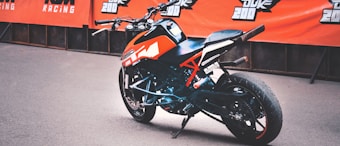 A sporty motorcycle with an orange and black color scheme is parked in front of a backdrop featuring branded banners. The design includes sharp angles and a modern aesthetic, suitable for racing or performance riding.