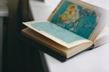 An open book with pages containing detailed maps, placed on a light-colored surface. The map on the visible page appears to show geographical regions in shades of blue, green, and brown.