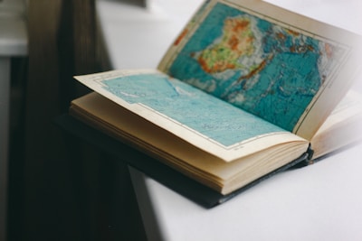 An open book with pages containing detailed maps, placed on a light-colored surface. The map on the visible page appears to show geographical regions in shades of blue, green, and brown.