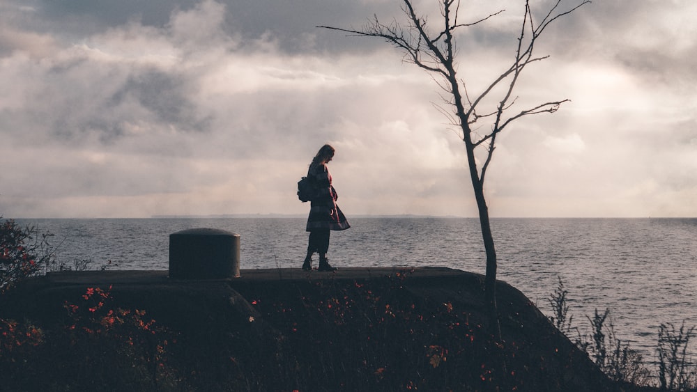 man in black jacket and black pants standing on rock near body of water during daytime