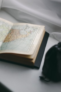 An open book displaying a detailed map with various regions and geographical features, lying on a white surface. The book's pages are slightly curled, indicating age or frequent use. Nearby, there is a dark, out-of-focus object, possibly a bag or a case.