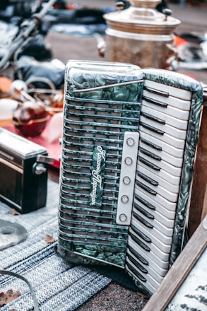 A vintage accordion with a pearl-like finish and black and white keys is prominently displayed on a textured surface. Surrounding it are various other items, including a brass samovar and what appears to be an old radio. The setting suggests a flea market or antique sale atmosphere.