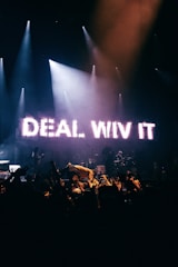 A concert stage is illuminated with bright lights, featuring a prominent LED sign displaying the words 'DEAL WIV IT'. The backdrop is dark, highlighting a crowd of people raising their hands. The atmosphere is energetic and lively, with musicians and a performer interacting with the audience.