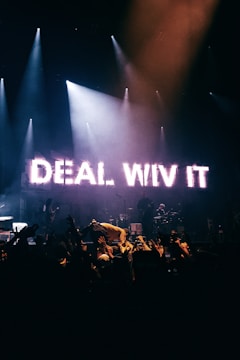 A concert stage is illuminated with bright lights, featuring a prominent LED sign displaying the words 'DEAL WIV IT'. The backdrop is dark, highlighting a crowd of people raising their hands. The atmosphere is energetic and lively, with musicians and a performer interacting with the audience.