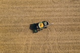 Aerial view of a green tractor harvesting a golden-brown field, with a trail indicating its movement. The field is uniformly covered with crop residue, and the tractor appears to be in operation, collecting grains.