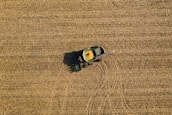 Aerial view of a green tractor harvesting a golden-brown field, with a trail indicating its movement. The field is uniformly covered with crop residue, and the tractor appears to be in operation, collecting grains.