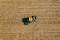 Aerial view of a green tractor harvesting a golden-brown field, with a trail indicating its movement. The field is uniformly covered with crop residue, and the tractor appears to be in operation, collecting grains.