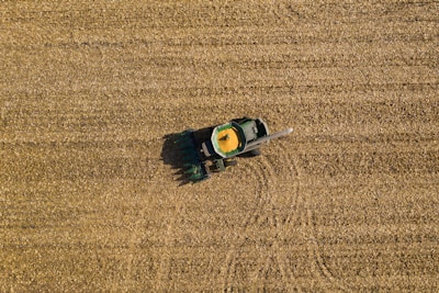 Aerial view of a green tractor harvesting a golden-brown field, with a trail indicating its movement. The field is uniformly covered with crop residue, and the tractor appears to be in operation, collecting grains.