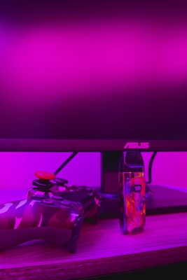 A gaming setup is featured with a camo-patterned game controller and a colorful vaporizer placed on a wooden desk. The background is illuminated with vibrant pink lighting, and a computer monitor with an ASUS logo is visible.