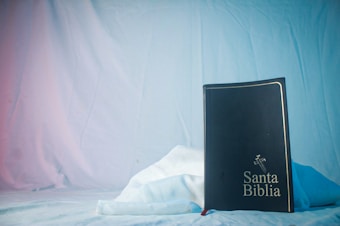 A black book with the title 'Santa Biblia' embossed on the cover is positioned upright against a soft, white fabric background. The book features a decorative cross design above the title. The backdrop is draped and softly lit with blue and pink hues.