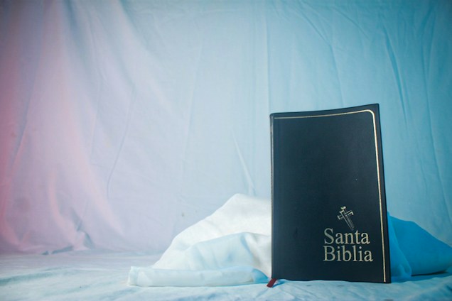 A black book with the title 'Santa Biblia' embossed on the cover is positioned upright against a soft, white fabric background. The book features a decorative cross design above the title. The backdrop is draped and softly lit with blue and pink hues.