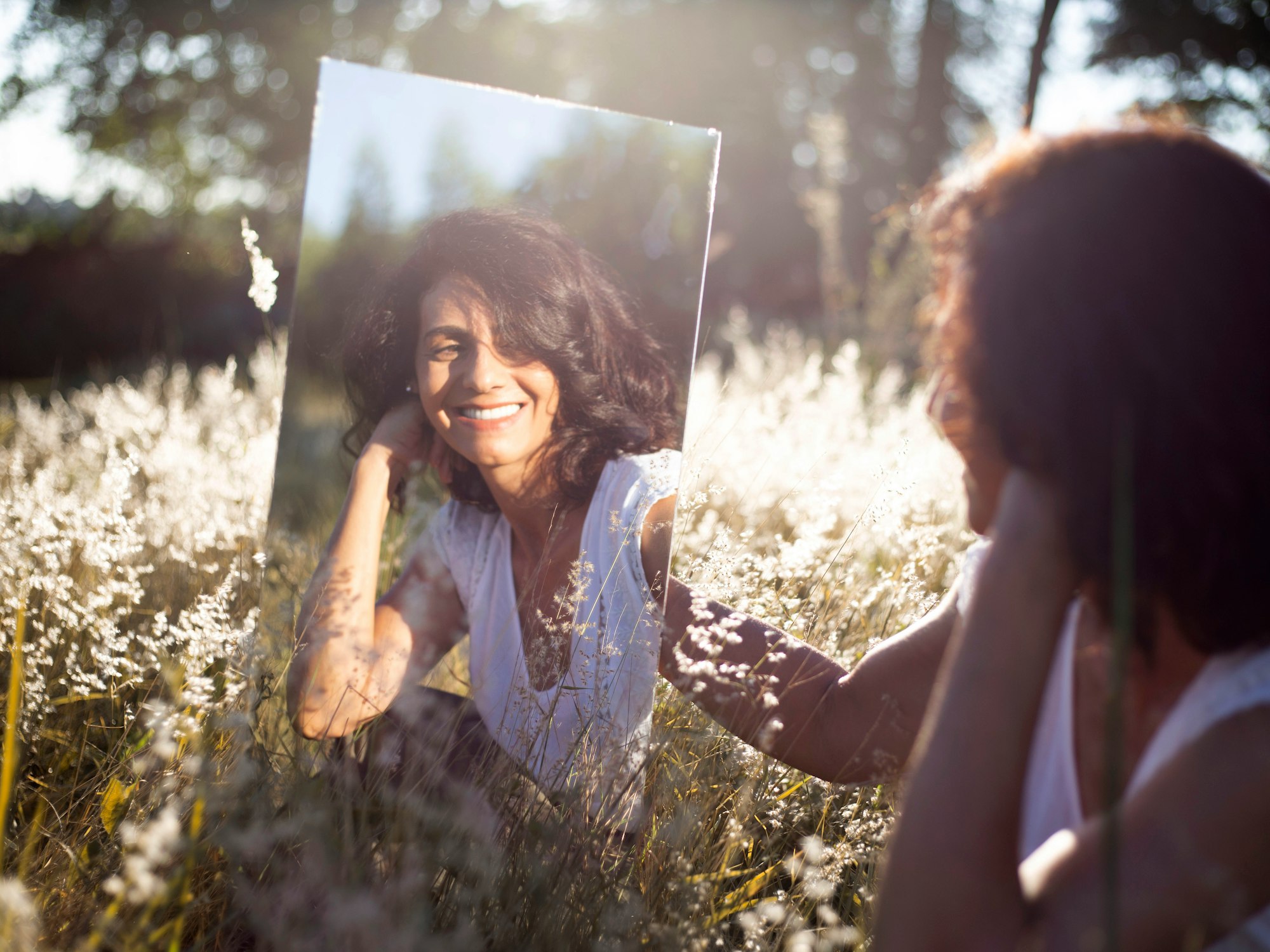 a smiling woman looking at herself in the mirror sitting in a sunny field in the nature