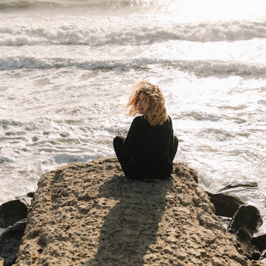 woman in black long sleeve shirt sitting on brown rock near sea during daytime