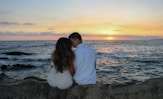 a man and a woman sitting on a rock near the ocean