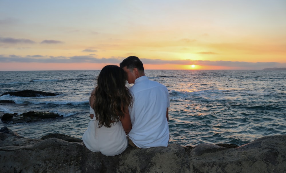 a man and a woman sitting on a rock near the ocean