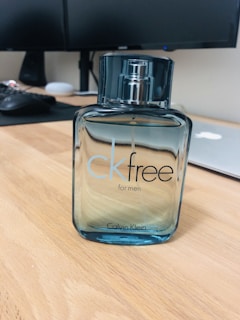 A bottle of CK Free for Men sits on a wooden desk, with a sleek and modern design. The bottle is transparent with a blue tint and has a metallic spray cap. In the background, there is a blurred office setting including a computer monitor, keyboard, and a closed laptop.