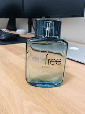A bottle of CK Free for Men sits on a wooden desk, with a sleek and modern design. The bottle is transparent with a blue tint and has a metallic spray cap. In the background, there is a blurred office setting including a computer monitor, keyboard, and a closed laptop.
